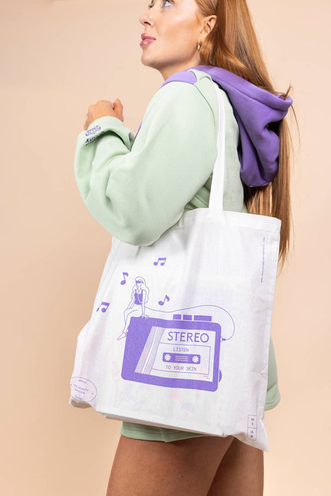 Woman holding a tote bag with a purple cassette design against a beige background